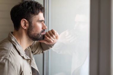 Bir adam pencerenin yanında duruyor, elini yüzüne koyup dışarı bakıyor. Önündeki manzarayı izlerken derin düşüncelere dalmış görünüyor..