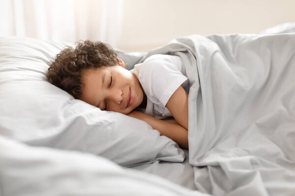 African American young boy peacefully sleeps in a bed, with his body curled up and his eyes closed. The room is dimly lit, and a blanket covers him.
