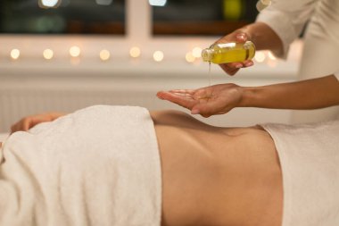 In a serene spa setting with ambient lighting, a professional masseuse is captured preparing for a soothing massage session by pouring massage oil onto their hands while the client waits clipart