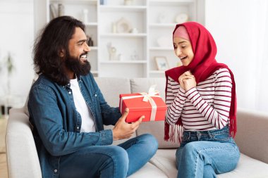 Doğulu bir adam ve bir kadın bir kanepede oturuyor, ellerinde hediye var. Konforlu bir oturma odasında, hediye verme ve kutlama anlarını paylaşıyor gibiler..