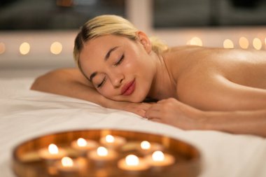 Yatakta uzanmış bir kadın var. SPA 'da etrafına mumlar dizilmiş. Oda loş bir şekilde aydınlatılmış, sıcak ve samimi bir atmosfer yaratıyor..