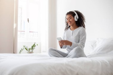 Genç bir İspanyol kadın yatağında bağdaş kurmuş, beyaz pijama ve kablosuz kulaklık takıyor, akıllı telefonuyla meşgul, muhtemelen bir şarkı seçiyor ya da bir mesaja cevap veriyor.
