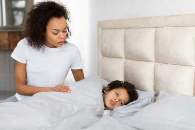 Endişeli bir Afro-Amerikan genç kadın bir yatağın yanında duruyor, nazikçe aşağı bakıyor, hasta gibi görünen ve rahat bir battaniyenin altında dinlenen bir çocuğa.