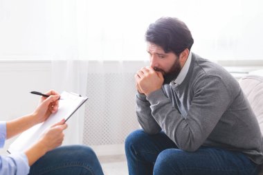 Sıkıntılı bir adam eli ağzında, sanki derin düşüncelere dalmış ya da endişeli bir şekilde not alan bir danışmanla konuşuyormuş gibi görülür. Birbirlerinin karşısında iyi aydınlatılmış bir ofiste oturuyorlar.