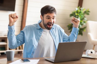 Sıradan mavi gömlekli neşeli bir adam yumruklarını kaldırıyor. Dizüstü bilgisayarının önünde. Yüzü sevinç ifade ediyor..