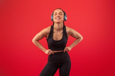 Neşeli bir kadın, siyah bir atlet ve tayt giymiş, canlı kırmızı bir arkaplan karşısında duruyor, kulaklıklarıyla müziğin keyfini çıkarırken, elleri kalçasında oynak poz veriyor..