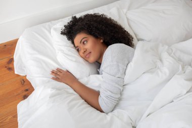 Mutlu İspanyol kadın, yumuşak beyaz bir yorganın altında, rahat bir şekilde yatakta yatıyor. Gözleri açık ve huzur içinde dinleniyor gibi görünüyor..