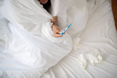 Cropped of woman appears to be sick, tucked under a white blanket, holding a thermometer to check her temperature. Tissues are scattered around her, suggesting she might have a cold or flu clipart