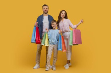 Üç kişilik neşeli bir aile, bir baba, bir anne ve küçük oğullarından oluşan, her biri çeşitli renkli alışveriş torbaları taşıyan, parlak sarı bir arka plana karşı dururlar.