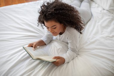 İspanyol kadın bir yatakta rahat bir şekilde uzanıyor, okuduğu bir kitaba dalmış. Önündeki metne odaklanmış ve rahatlamış görünüyor..