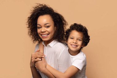 Kıvırcık saçlı neşeli bir Afrikalı Amerikalı anne genç oğlunu sevgiyle kucaklıyor ve her ikisi de geniş bir şekilde gülümsüyor. Tarafsız bir bej arka plana karşı poz veriyorlar, yakın plan.
