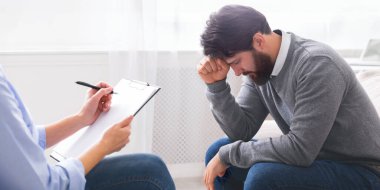 Bir adam sandalyeye oturtuluyor, başı ellerinde, stres veya hayal kırıklığı belirtileri gösteriyor, derin düşüncelere dalmış, terapistle seans sırasında stres veya tefekküre dalmış.