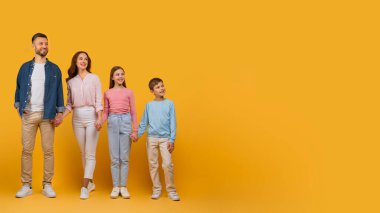 İki yetişkin ve iki çocuktan oluşan mutlu bir aile yan yana durur, gülümser ve sollarına bakarlar, canlı sarı bir zemin karşısında, boş alana bakarlar.