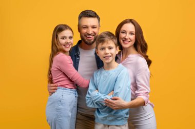 Bir baba, anne, kız ve oğuldan oluşan birbirine yakın bir aile, kameraya doğru gülümserken birbirlerine yakın duruyorlar. Mutlu ve mutlu görünüyorlar.