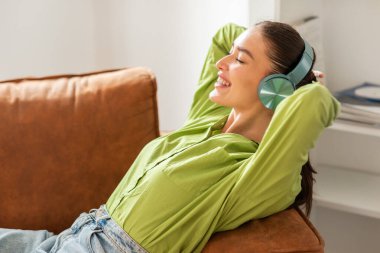 Bir kadın kanepede oturur, kulaklıkla müzik dinlemeye dalmıştır. Rahatlamış ve odaklanmış görünüyor. Müzikten zevk alıyor..