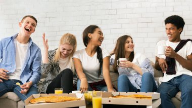 Modern bir koltukta oturan çeşitli arkadaş grupları pizza dilimlerinin tadını çıkarırken canlı sohbetler yapıyorlar. Bu sıradan ve keyifli anı paylaşırken kahkahalar odayı dolduruyor..