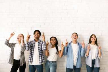 Çok ırklı beş gençten oluşan çeşitli bir grup beyaz tuğla bir duvarın önünde yan yana duruyor, hepsi gülümsüyor ve yukarı bakarken aynı zamanda işaret parmaklarıyla işaret ediyorlar.