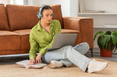 Odaklanmış genç bir kadın sırtı kanepeye yaslanmış, kulaklık takmış ve dizüstü bilgisayar kullanıyor, muhtemelen uzaktan çalışma ya da çalışmayla meşgul.