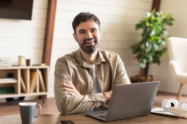 Neşeli bir adam, iyi ışıklandırılmış ev ofisinin ahşap masasında oturuyor. Kollarını dizüstü bilgisayarının önünde kavuşturuyor. Kendine güvenen bir gülümsemeyle ya işe ara veriyor ya da video konferansına katılıyor.