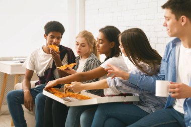 Bir grup etnik çeşitliliğe sahip genç, açık bir kutudan büyük bir pizzayı paylaşıyor. Parlak bir odada birlikte güzel vakit geçiriyorlar..