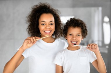 Her ikisi de beyaz tişörtler giyen Afrikalı Amerikalı anne ve küçük çocuğu, dişlerini iyi aydınlatılmış bir banyoda fırçalarken neşeli bir anı paylaşıyorlar.