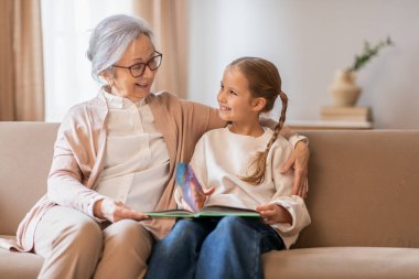 Gri saçlı yaşlı bir kadın rahat bir kanepede genç bir kızın yanında oturuyor. Her ikisi de bir masal kitabı okurken içten bir şekilde gülümsüyor ve bir bağ kurma anını paylaşıyorlar.