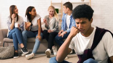 Siyahi genç çocuk ön planda görünür bir şekilde stresli ve rahatsız görünüyor. Arkadaşlarından oluşan bir grup, iyi aydınlatılmış bir odada kanepede oturmuş neşeli bir sohbete ve kahkahaya dalmışken.