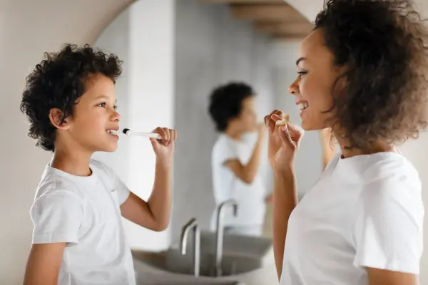 stock image African American woman is brushing her teeth next to a little boy. They are standing in front of a bathroom sink. The woman holds a toothbrush while the little boy watches intently.