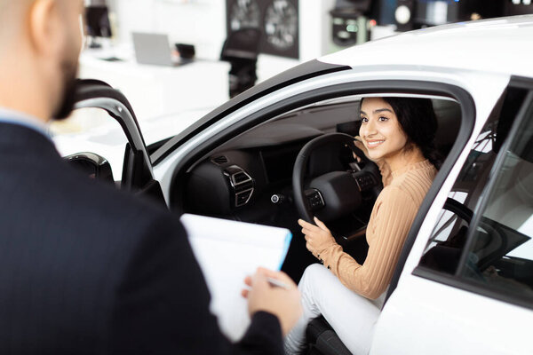 A young Indian woman sits behind the wheel of a new car, smiling at the camera with a sense of excitement. She is engaging in a test drive at an automotive dealership showroom