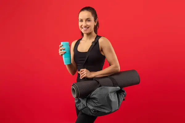 stock image A woman is standing while holding a yoga mat in one hand and a cup in the other hand. She appears to be ready for a yoga session or workout, balancing the items with ease.