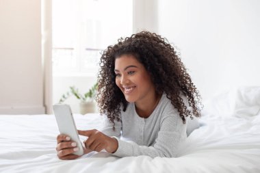 Kıvırcık saçlı neşeli, genç bir İspanyol kadın beyaz bir yatakta karnının üzerinde uzanmış, akıllı telefonuyla meşgul. Rahatlamış ve mutlu görünüyor, muhtemelen internette geziniyor ya da sohbet ediyor.