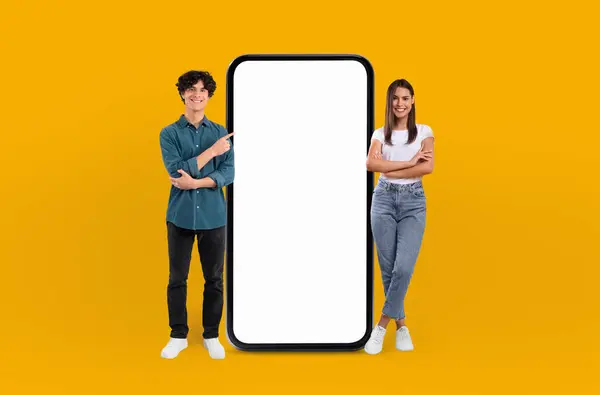 stock image A man and a woman are standing side by side next to a large phone. The man is holding a notepad and pen while the woman looks at the phone screen. They appear to be discussing something important.