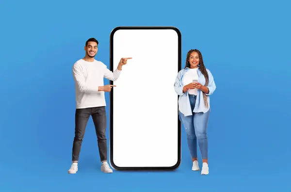 Multiethnic man and woman are standing side by side next to large phone, looking at the screen and appearing to have conversation. The phone is significantly bigger than them, emphasizing its size.