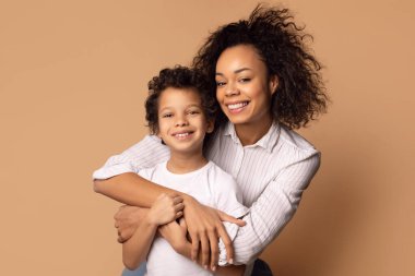 Neşeli bir Afrikalı Amerikalı anne, mutluluk saçan kıvırcık saçlı genç oğluna sıkıca sarılıyor. Her ikisi de samimi bir şekilde gülümsüyor, çünkü aralarında özel bir bağ ve sevgi hissediyorlar.