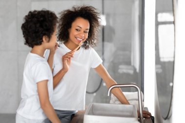 Siyahi bir kadın, dişlerini fırçalayan genç bir çocuğun yanında görüldü. Anne bir diş fırçası tutuyor ve aynadaki yansımasına bakıyor. Oğul onun hareketlerini taklit ediyor.