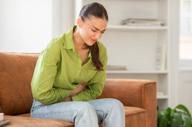 Bir kadın kanepeye oturmuş, bir eli karnında dinleniyor. Rahatsızlık ya da acı çekiyor gibi görünüyor, muhtemelen mide krampları ya da hazımsızlık çekiyor..