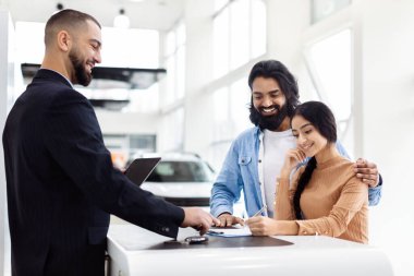 Genç Hintli bir çift, yeni bir araba satın alma sürecinde. Parlak bir araba galerisinde, dost canlısı bir satıcının sunduğu belgeleri imzalıyorlar.