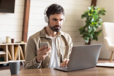 Bir adam ahşap bir masada oturuyor, kulaklık takarken dizüstü bilgisayarıyla dikkatle meşgul oluyor. Aynı anda sağ elinde bir akıllı telefon tutuyor.