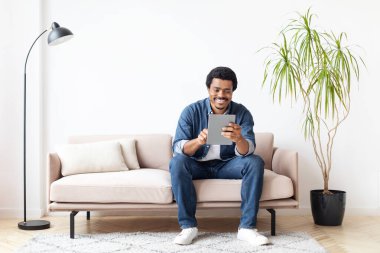 Afrikalı Amerikalı bir adam bir koltuğa oturmuş, bu kapalı ortamda bir tablet cihazla meşgul, ev ortamında modern teknolojiyi ve eğlenceyi sergiliyor..