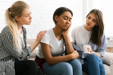 Üç çok ırklı kız bir kanepede yakın otururken, bir tanesi sıkıntılı görünüyor, diğer ikisi ise rahat görünüyor. Oda aydınlık ve destekleyici bir ortam hissi uyandırıyor.