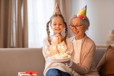 An older woman and a young girl sit on a couch, with a birthday cake in front of them. The woman is lighting the candles on the cake while the girl eagerly watches clipart