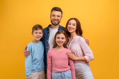 İki yetişkin ve iki çocuktan oluşan dört kişilik bir aile, canlı sarı bir arkaplan önünde bir fotoğrafa gülümseyen ve poz veren ebeveynler çocukların ellerini tutarken bir arada duruyorlar.
