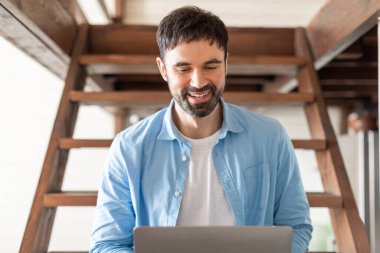 Sakallı neşeli genç bir adam, kapalı alanda çalışırken dizüstü bilgisayarına odaklanır. İyi ışıklandırılmış, havadar bir yerde, ahşap bir merdivende oturur. Bu da bir ev ya da sıradan bir ofis ortamı olduğunu gösterir..