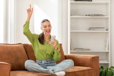 Bronz deri koltukta bağdaş kurmuş bir kadın muzaffer bir ifadeyle oturuyor ve kolunu zafer jesti ile kaldırıyor. Diğer elinde bir akıllı telefon var, muhtemelen iyi haberler almıştır.