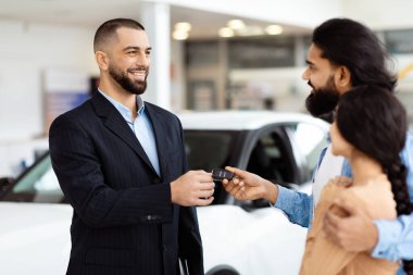İyi giyimli bir satıcı, araba galerisinde neşeli bir Hintli çifte araba anahtarlarını sunuyor.