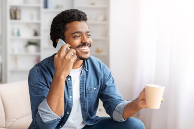 Siyah adam kapalı bir koltukta oturuyor, cep telefonuyla konuşuyor. Odaklanmış görünüyor, telefonu kulağına dayayıp konuşuyor. Arka plan tipik bir oturma odası düzenini gösteriyor..