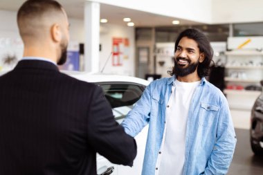 Two men are standing in a car showroom, shaking hands in agreement. The men appear serious and focused on finalizing a deal for a car purchase clipart