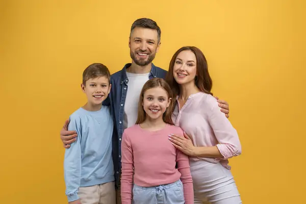 stock image A family of four, consisting of two adults and two children, stands together in front of a vibrant yellow background, smiling and posing for a photograph, with the parents holding the children hands
