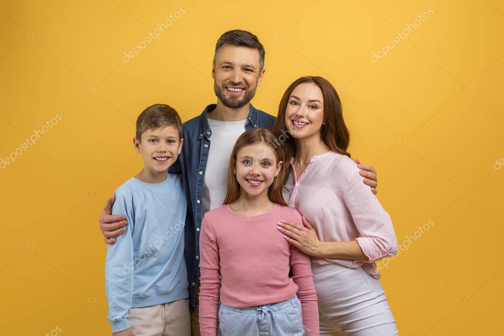 A family of four, consisting of two adults and two children, stands together in front of a vibrant yellow background, smiling and posing for a photograph, with the parents holding the children hands