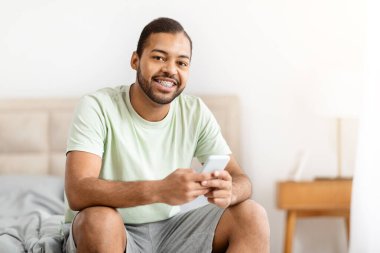 Neşeli, Afro-Amerikan bir adam rahat bir duruş içinde yatağında oturmuş elinde akıllı bir telefon tutuyor..